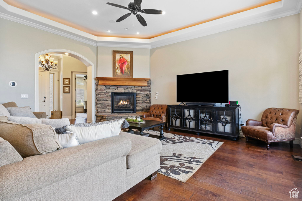 Living room featuring crown molding, dark wood-type flooring, ceiling fan with notable chandelier, a tray ceiling, and a stone fireplace