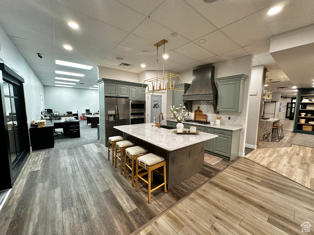 Kitchen with appliances with stainless steel finishes, an island with sink, wood-type flooring, a drop ceiling, and custom exhaust hood