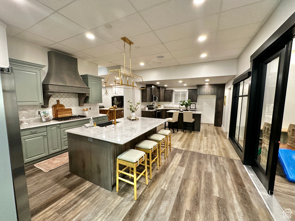 Kitchen with a paneled ceiling, a center island with sink, premium range hood, hardwood / wood-style flooring, and tasteful backsplash