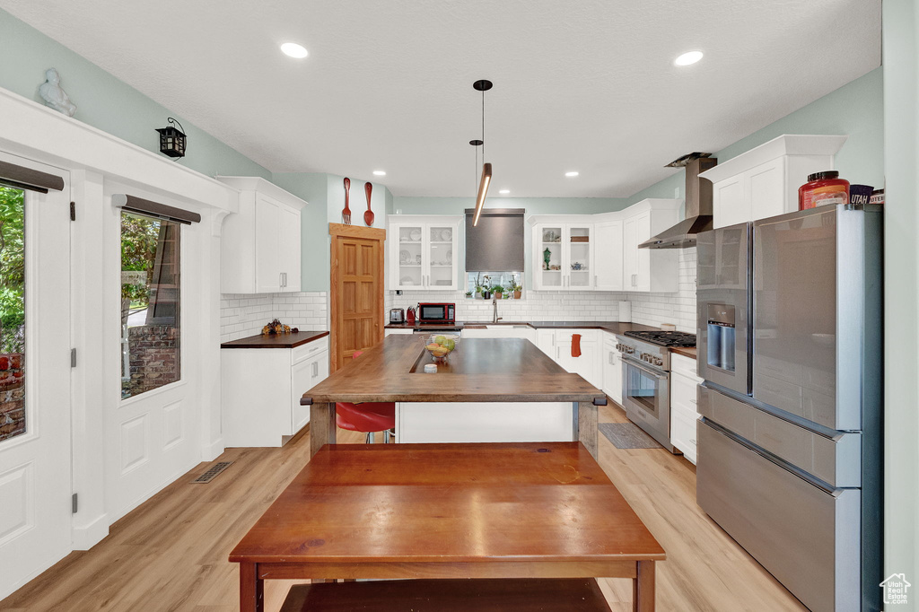 Kitchen featuring tasteful backsplash, light wood-type flooring, wall chimney range hood, white cabinets, and appliances with stainless steel finishes