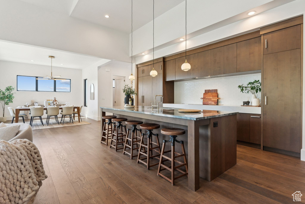 Kitchen featuring an island with sink, hanging light fixtures, a breakfast bar, dark wood-type flooring, and dark stone counters