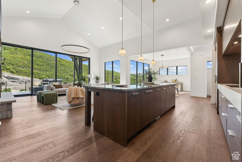 Kitchen with decorative light fixtures, a kitchen island with sink, high vaulted ceiling, hardwood / wood-style flooring, and dark brown cabinetry