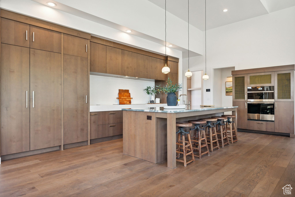 Kitchen featuring stainless steel double oven, an island with sink, hanging light fixtures, a towering ceiling, and dark wood-type flooring