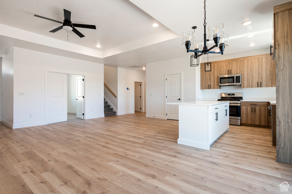 Kitchen with light wood-type flooring, ceiling fan with notable chandelier, a raised ceiling, stainless steel appliances, and pendant lighting