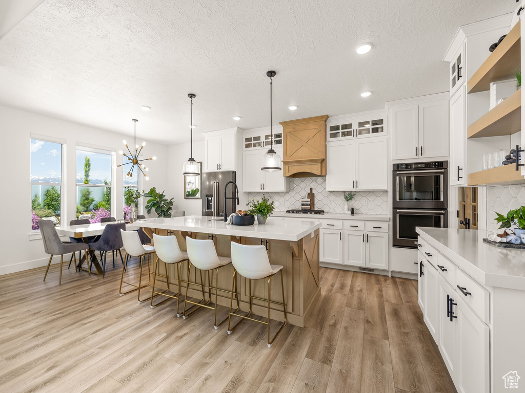 Kitchen featuring light hardwood / wood-style floors, tasteful backsplash, a center island with sink, stainless steel appliances, and decorative light fixtures