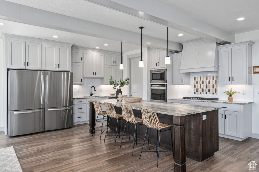 Kitchen with custom exhaust hood, stainless steel appliances, wood-type flooring, hanging light fixtures, and white cabinets