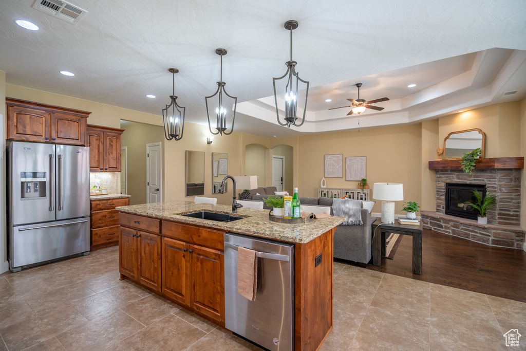 Kitchen featuring a stone fireplace, a center island with sink, a raised ceiling, sink, and appliances with stainless steel finishes