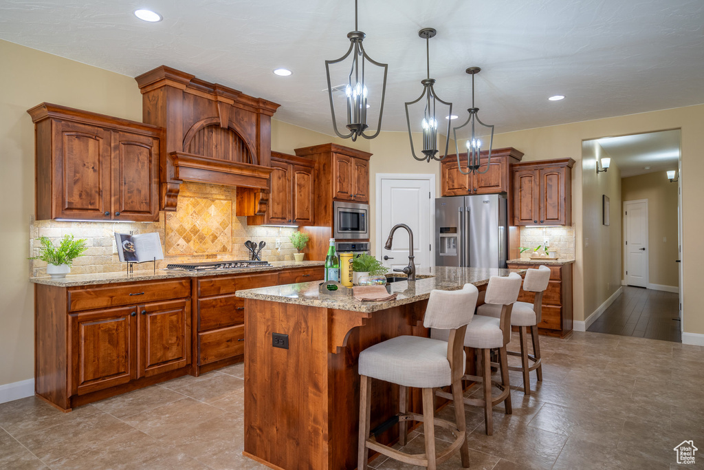 Kitchen with an island with sink, stainless steel appliances, pendant lighting, light stone counters, and backsplash