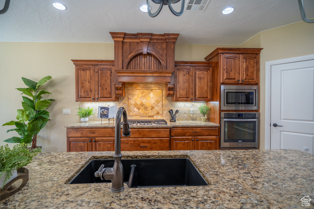 Kitchen with backsplash, custom exhaust hood, light stone countertops, appliances with stainless steel finishes, and sink