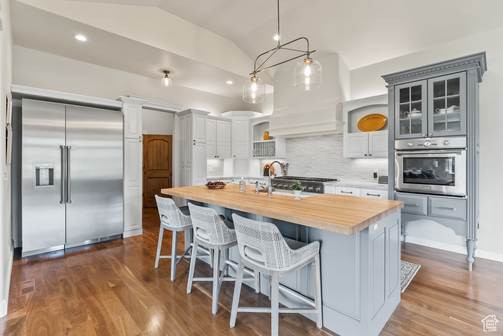 Kitchen with tasteful backsplash, butcher block countertops, an island with sink, vaulted ceiling, and appliances with stainless steel finishes