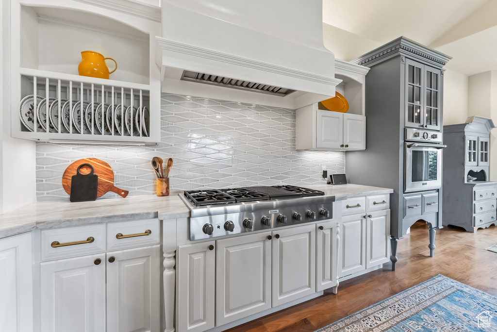 Kitchen with light stone counters, tasteful backsplash, hardwood / wood-style floors, appliances with stainless steel finishes, and premium range hood