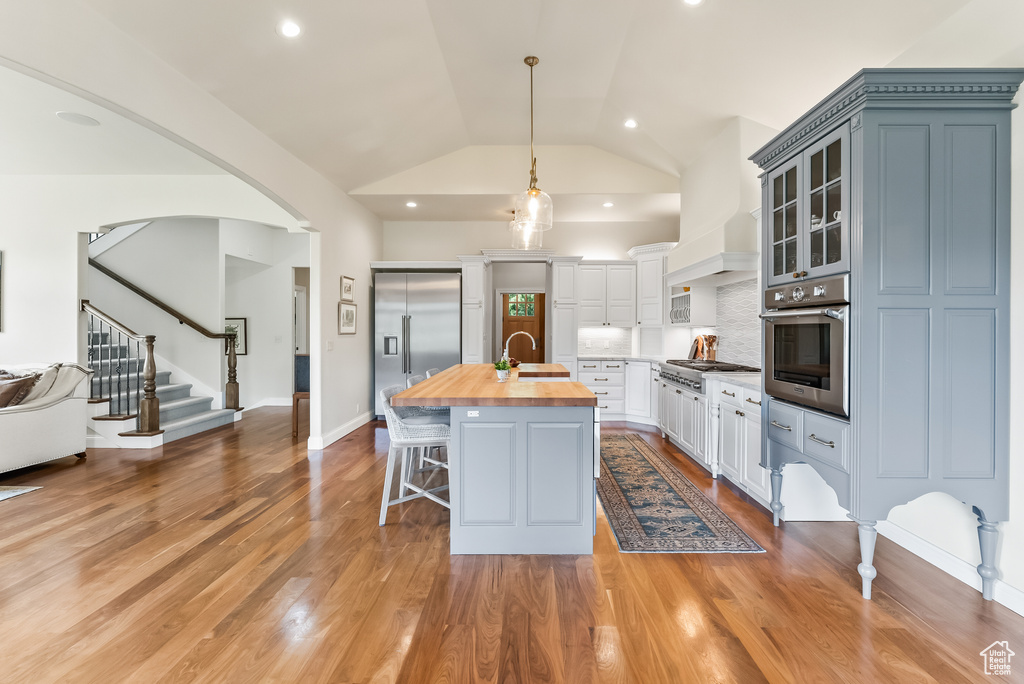 Kitchen with hanging light fixtures, stainless steel appliances, butcher block countertops, a kitchen island with sink, and wood-type flooring