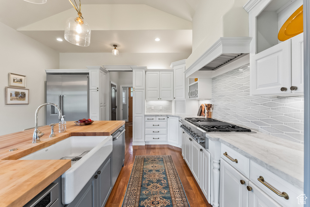 Kitchen with white cabinetry, custom range hood, appliances with stainless steel finishes, decorative light fixtures, and tasteful backsplash