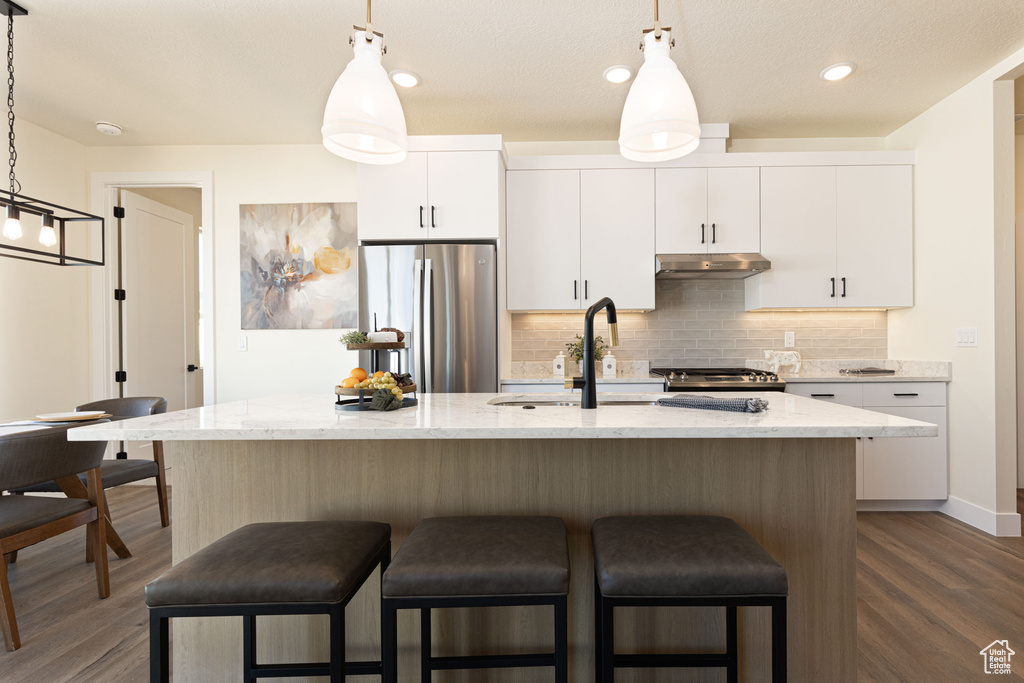 Kitchen with decorative light fixtures, dark wood-type flooring, an island with sink, stainless steel refrigerator, and white cabinetry