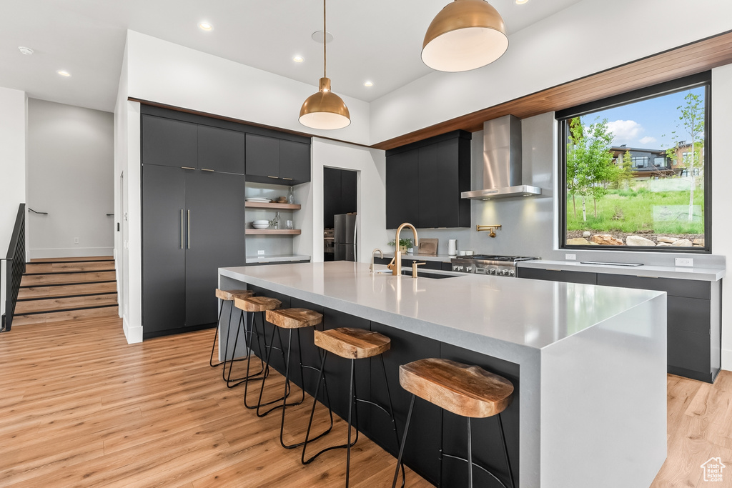 Kitchen featuring hanging light fixtures, light hardwood / wood-style floors, a center island with sink, wall chimney range hood, and sink