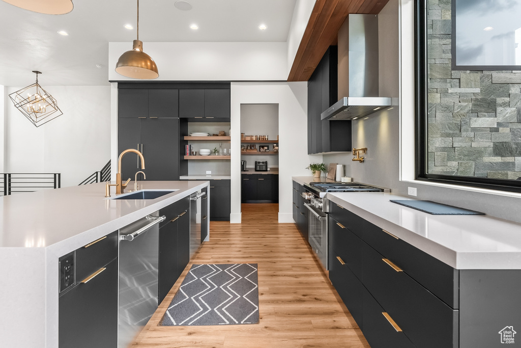 Kitchen featuring wall chimney range hood, hanging light fixtures, light hardwood / wood-style floors, stainless steel appliances, and a kitchen island with sink