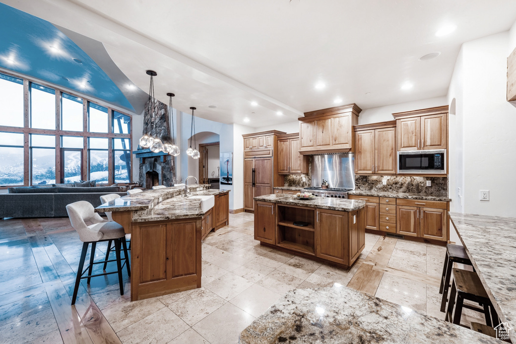 Kitchen with light stone counters, backsplash, hanging light fixtures, built in appliances, and a kitchen island with sink