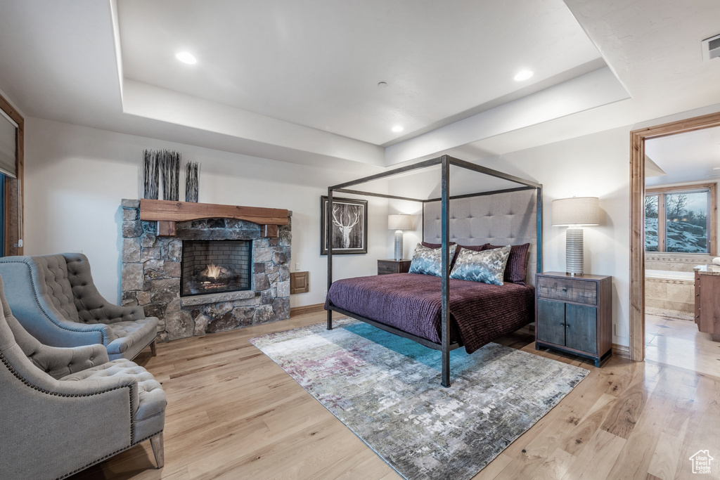 Bedroom with a stone fireplace, a tray ceiling, ensuite bathroom, and light hardwood / wood-style flooring