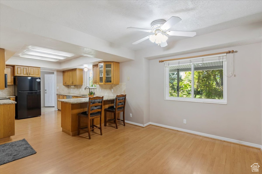 Kitchen featuring light hardwood / wood-style floors, kitchen peninsula, backsplash, black refrigerator, and a breakfast bar