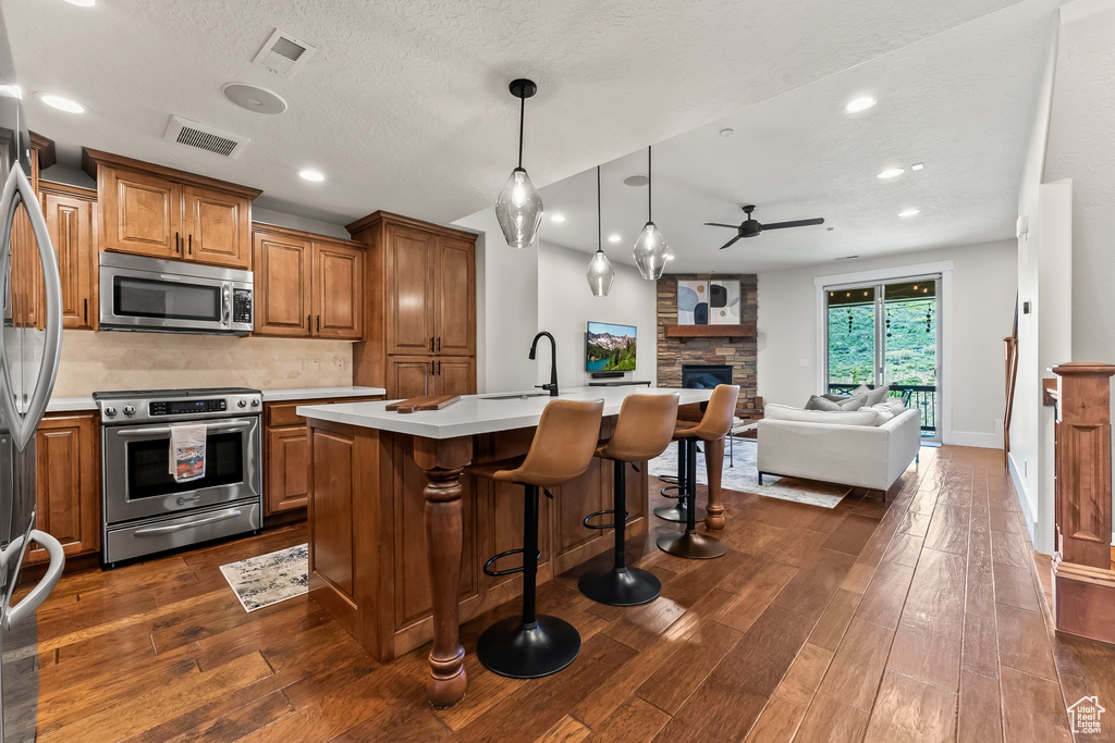 Kitchen featuring dark hardwood / wood-style flooring, a fireplace, a center island with sink, ceiling fan, and appliances with stainless steel finishes