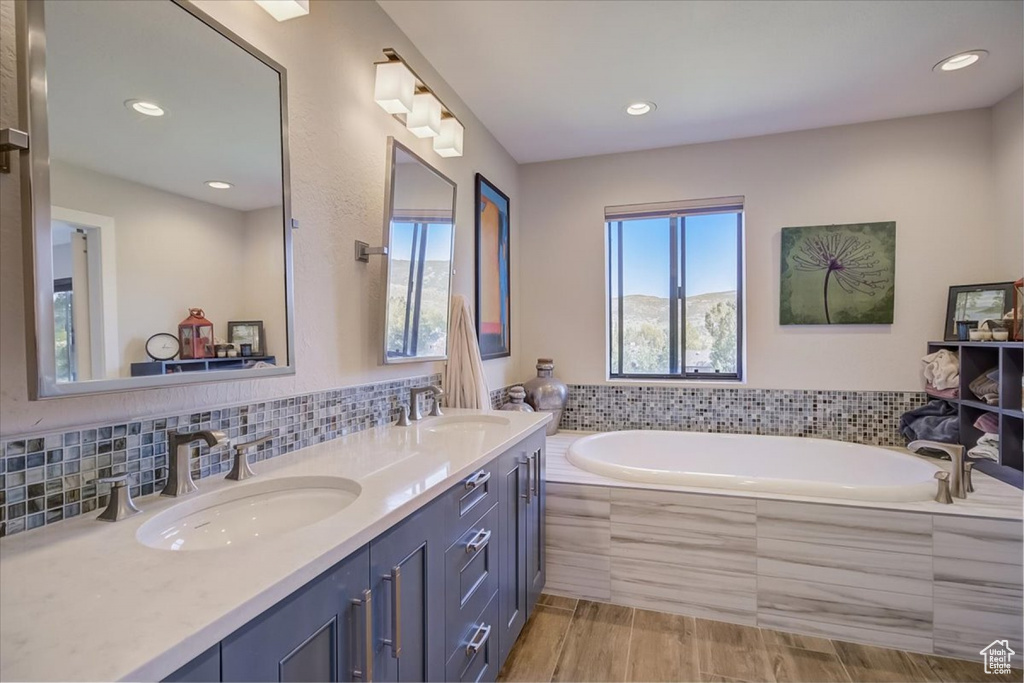 Bathroom with dual sinks, vanity with extensive cabinet space, tiled tub, and backsplash