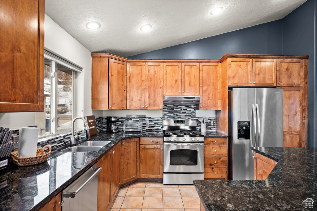 Kitchen with dark stone counters, tasteful backsplash, stainless steel appliances, light tile floors, and lofted ceiling