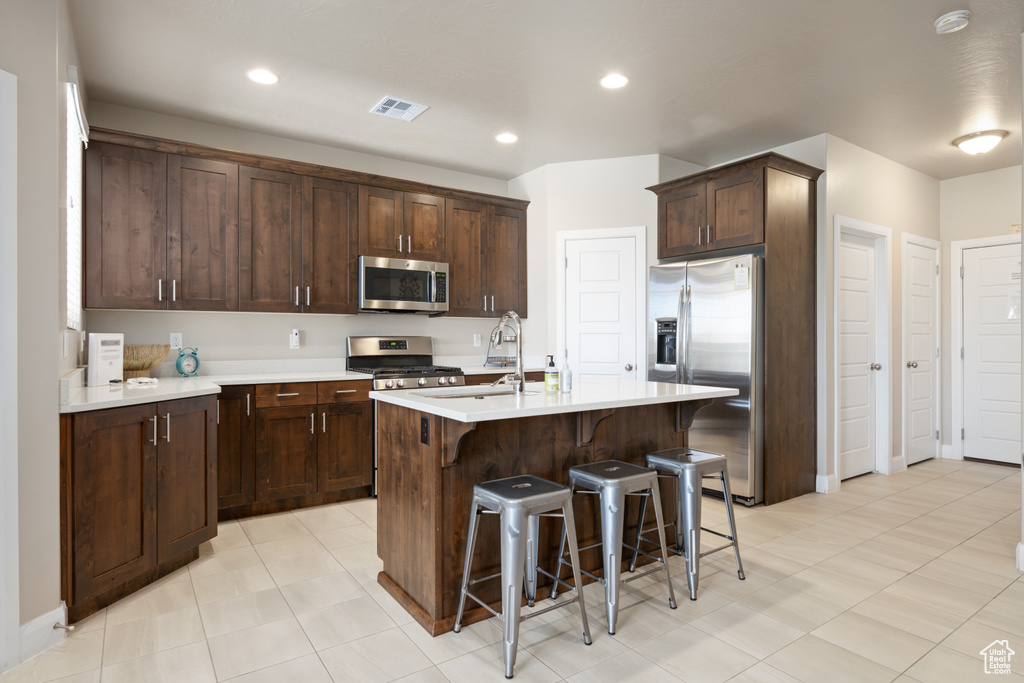 Kitchen featuring a kitchen breakfast bar, an island with sink, stainless steel appliances, light tile flooring, and dark brown cabinetry