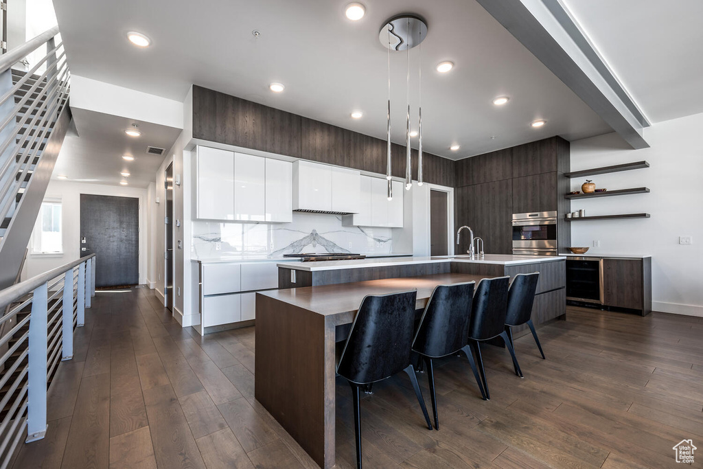 Kitchen with dark hardwood / wood-style floors, oven, hanging light fixtures, an island with sink, and white cabinets