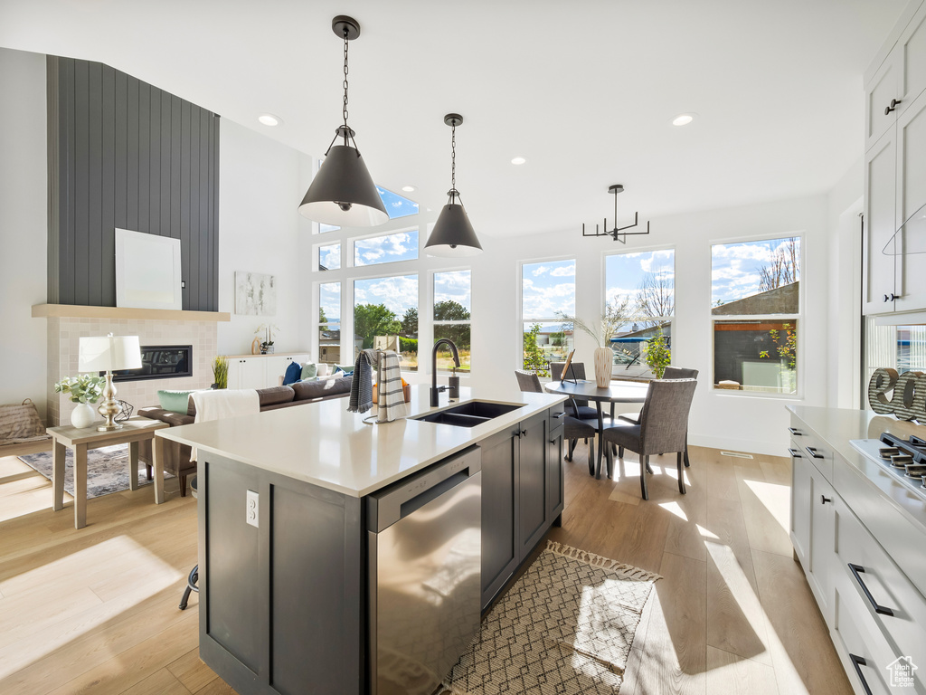 Kitchen with light wood-type flooring, a kitchen island with sink, dishwasher, sink, and decorative light fixtures