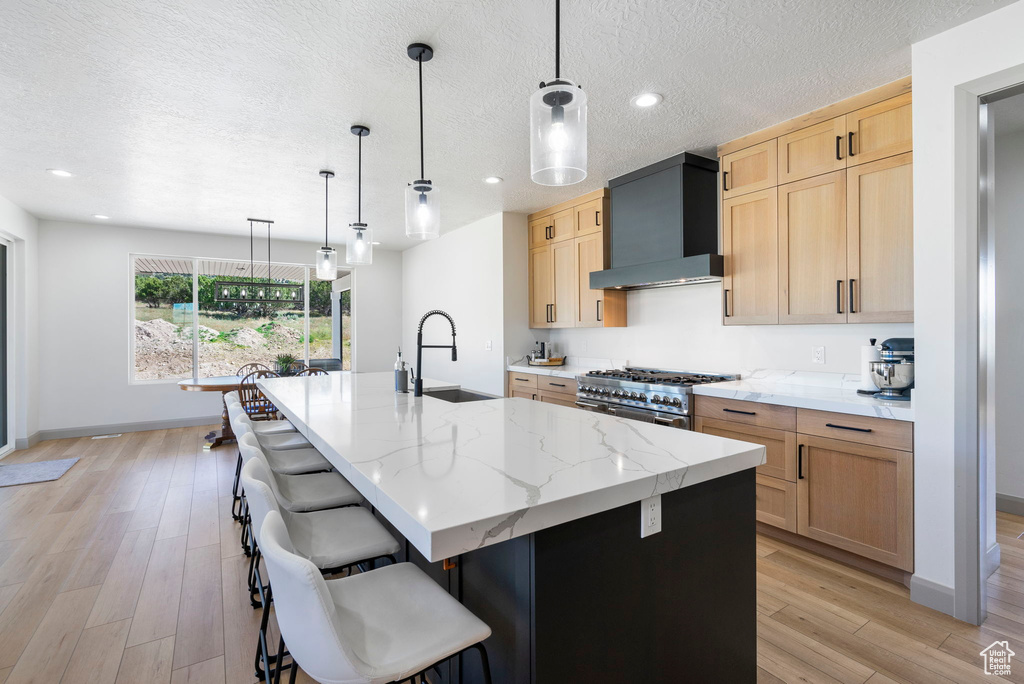 Kitchen featuring custom exhaust hood, light stone countertops, an island with sink, light wood-type flooring, and double oven range