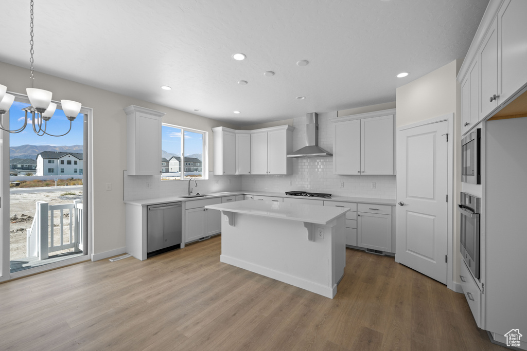 Kitchen featuring wall chimney exhaust hood, white cabinetry, appliances with stainless steel finishes, a center island, and light hardwood / wood-style floors