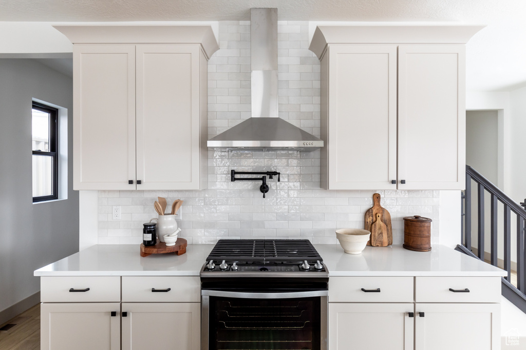 Kitchen featuring wall chimney exhaust hood, tasteful backsplash, white cabinetry, and stainless steel gas range oven