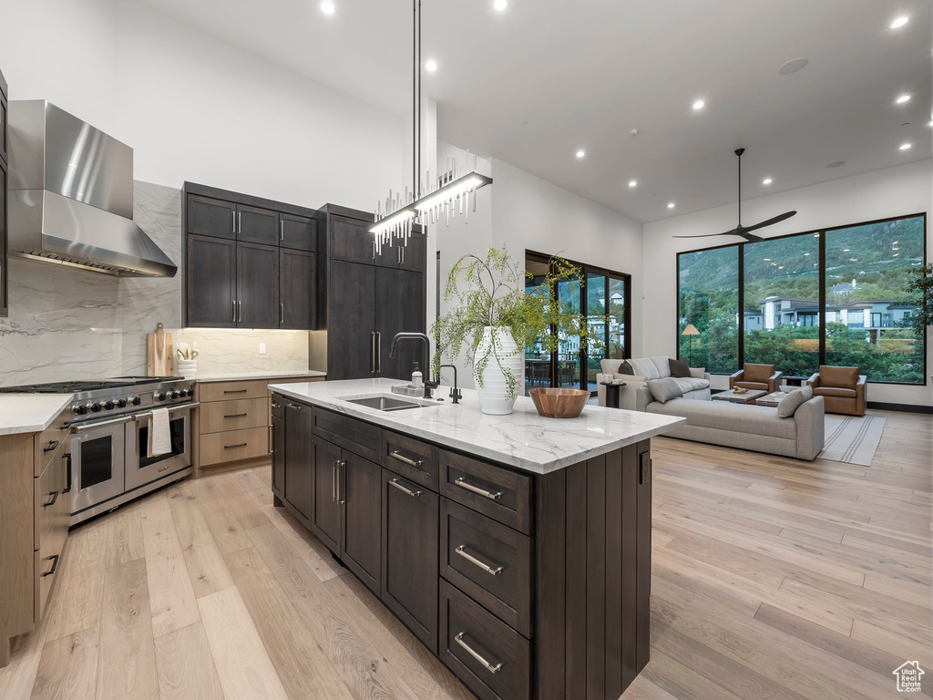 Kitchen with wall chimney range hood, light hardwood / wood-style flooring, a center island with sink, tasteful backsplash, and double oven range