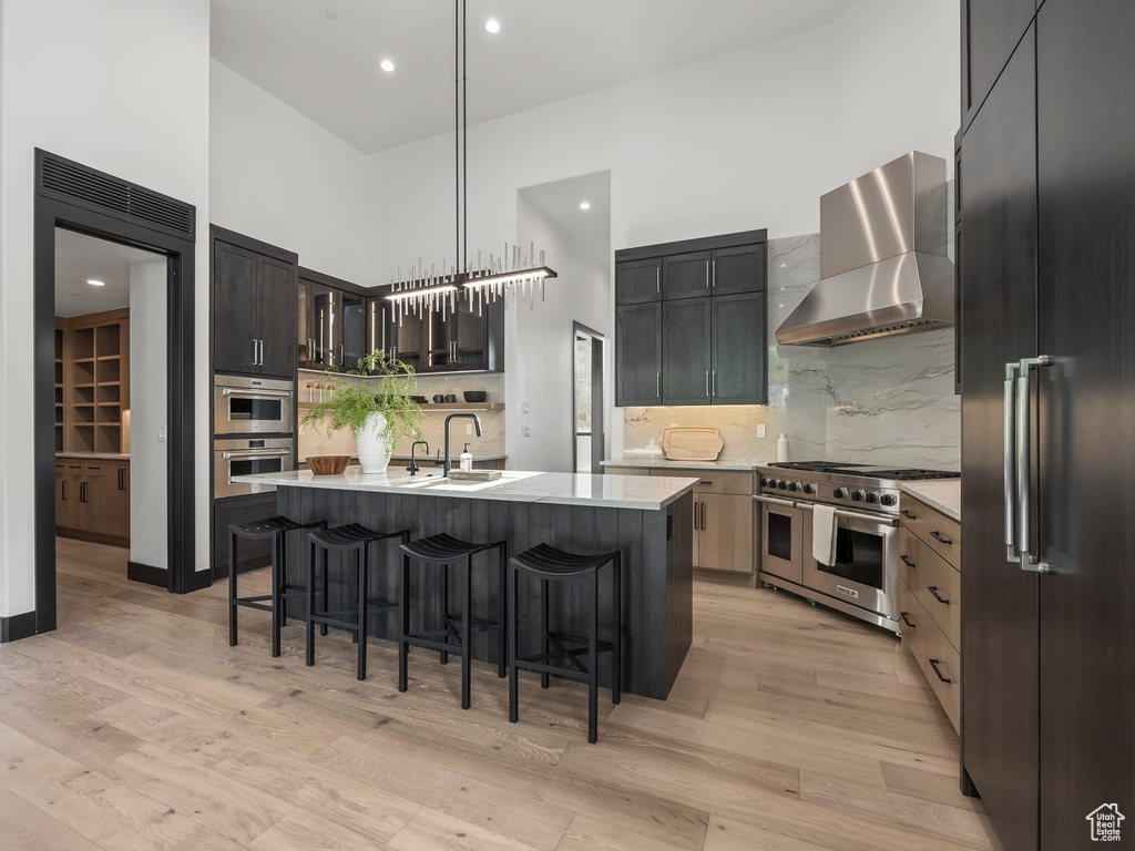 Kitchen with a towering ceiling, an island with sink, wall chimney exhaust hood, premium appliances, and light wood-type flooring