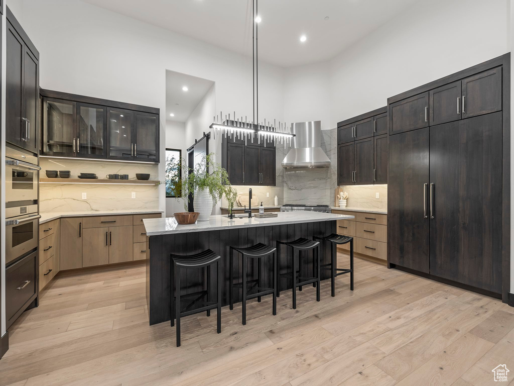 Kitchen featuring light hardwood / wood-style floors, hanging light fixtures, stainless steel double oven, a kitchen island with sink, and wall chimney exhaust hood