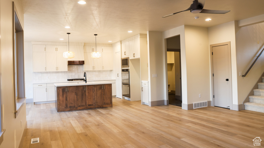 Kitchen with an island with sink, hanging light fixtures, light hardwood / wood-style flooring, backsplash, and white cabinets