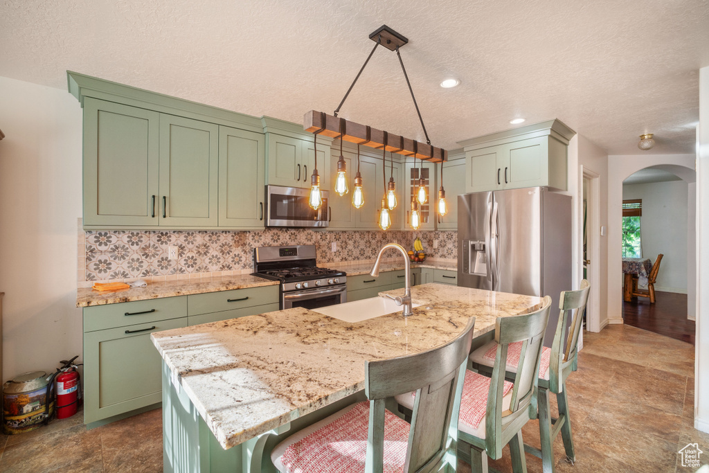 Kitchen featuring stainless steel appliances, hanging light fixtures, an island with sink, and green cabinets