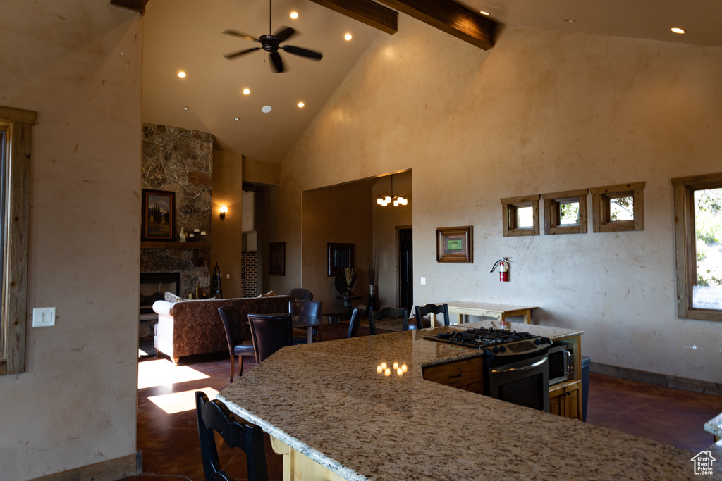 Kitchen with a stone fireplace, beamed ceiling, ceiling fan with notable chandelier, high vaulted ceiling, and gas range