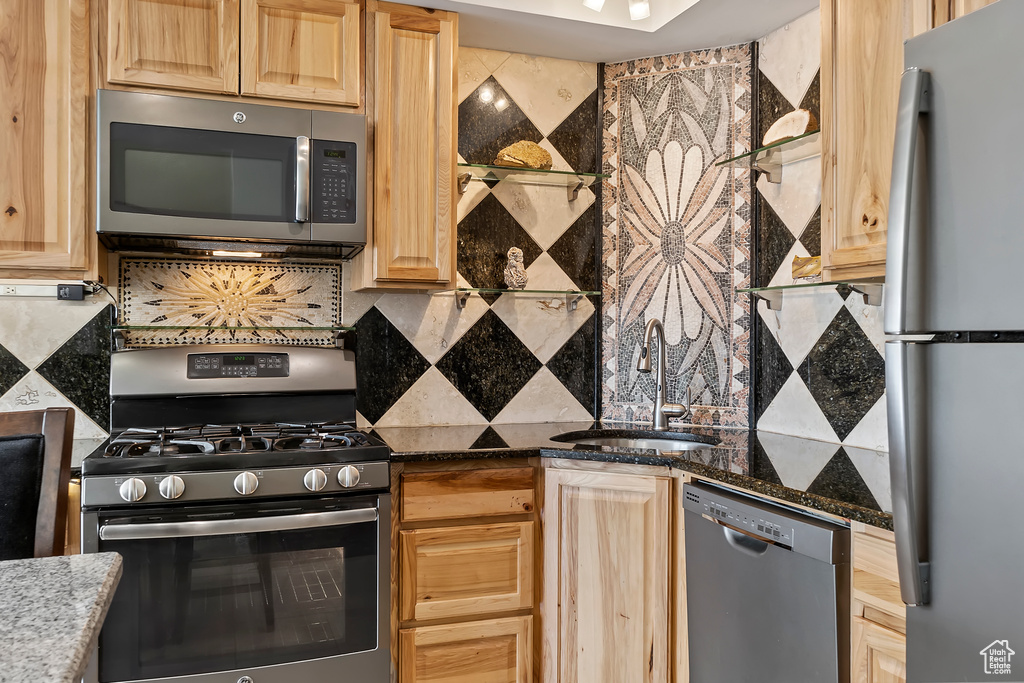 Kitchen with dark stone countertops, stainless steel appliances, backsplash, sink, and light brown cabinetry