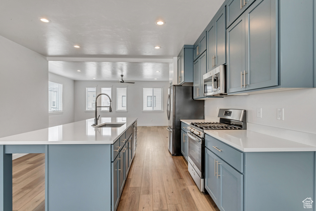 Kitchen with light hardwood / wood-style flooring, appliances with stainless steel finishes, a kitchen island with sink, sink, and ceiling fan