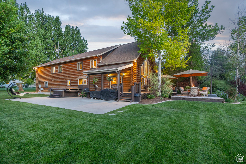 Back of property at dusk with a hot tub, a patio, log siding, and a lawn