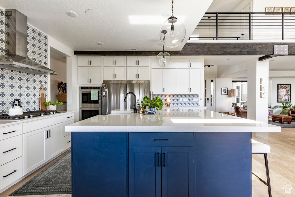 Kitchen featuring light hardwood / wood-style floors, backsplash, wall chimney exhaust hood, a kitchen island with sink, and appliances with stainless steel finishes