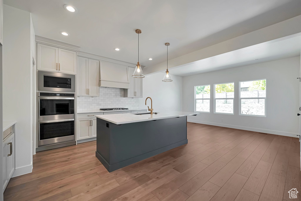 Kitchen featuring custom range hood, light hardwood / wood-style flooring, appliances with stainless steel finishes, a kitchen island with sink, and tasteful backsplash