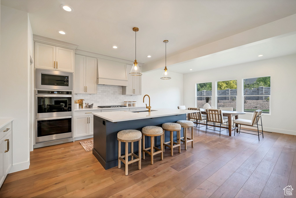 Kitchen with custom range hood, light hardwood / wood-style flooring, a kitchen island with sink, built in microwave, and decorative backsplash