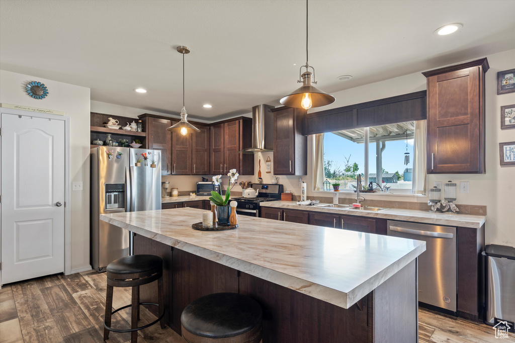 Kitchen with a center island, appliances with stainless steel finishes, hardwood / wood-style floors, wall chimney exhaust hood, and decorative light fixtures