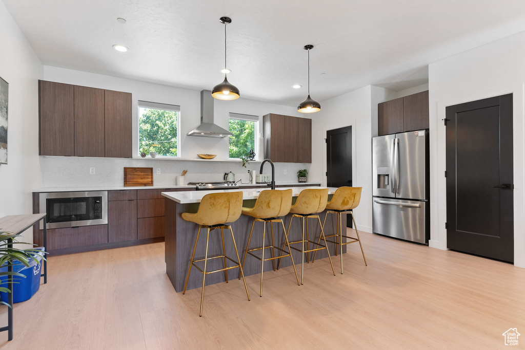 Kitchen featuring wall chimney exhaust hood, light hardwood / wood-style floors, stainless steel appliances, and a kitchen island with sink