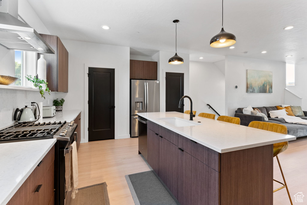 Kitchen with stainless steel appliances, light hardwood / wood-style flooring, a kitchen island with sink, wall chimney exhaust hood, and sink