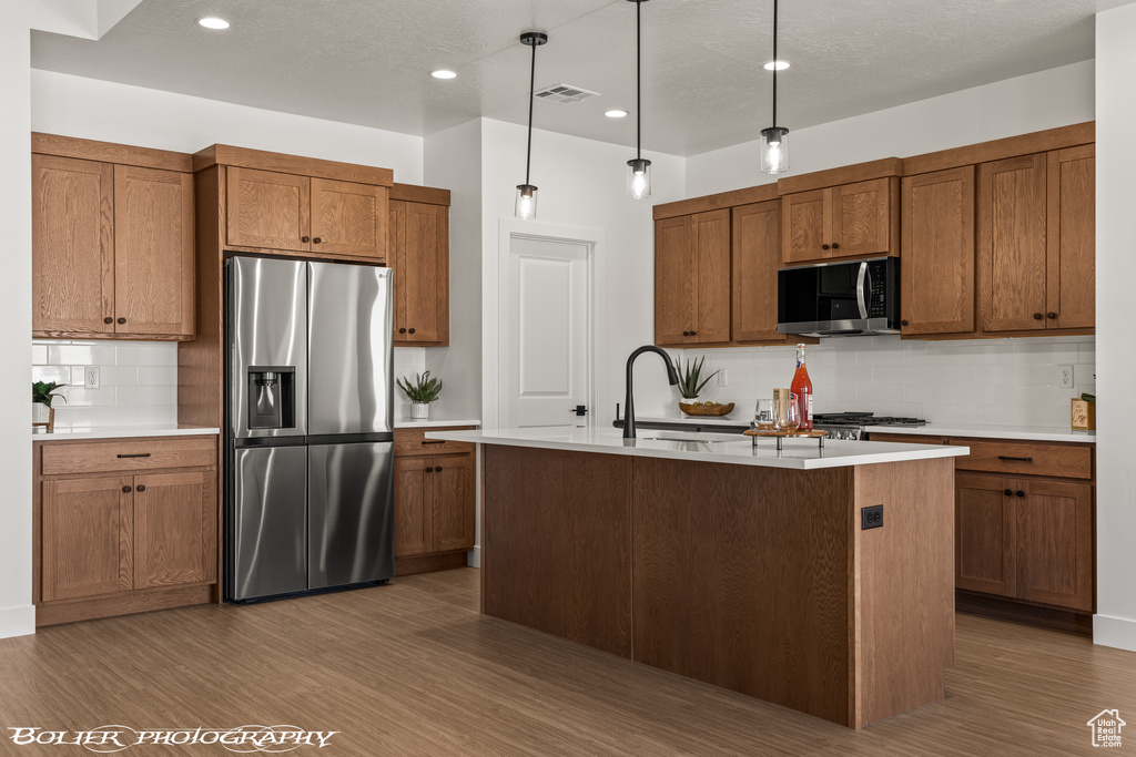 Kitchen with dark wood-type flooring, tasteful backsplash, an island with sink, stainless steel appliances, and decorative light fixtures