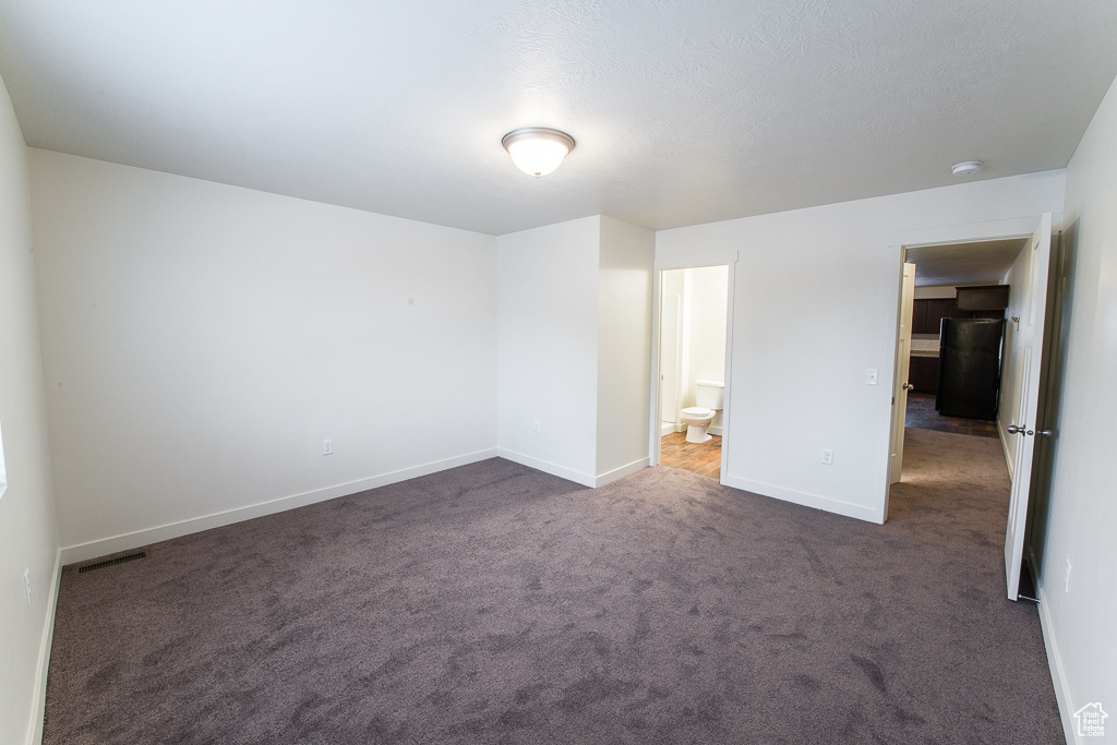 Unfurnished bedroom with black fridge, dark colored carpet, and connected bathroom