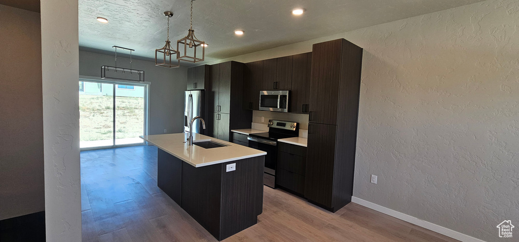 Kitchen with appliances with stainless steel finishes, sink, light hardwood / wood-style floors, decorative light fixtures, and a kitchen island with sink